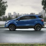 A blue 2021 Ford EcoSport is shown from the side driving on an open road past a city.