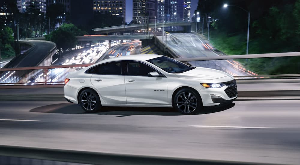 A white 2022 Chevy Malibu is shown from the side driving on an overpass after winning a 2022 Chevy Malibu vs 2022 Toyota Camry comparison.