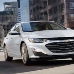 A white 2022 Chevy Malibu is shown from the front driving through a city.