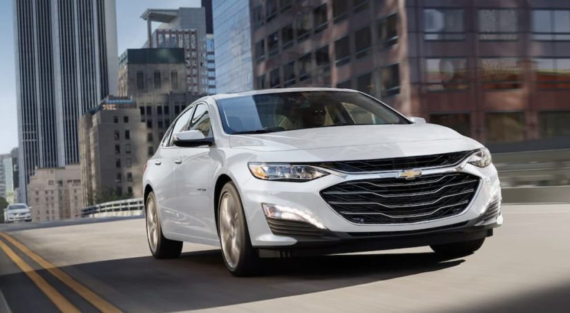 A white 2022 Chevy Malibu is shown from the front driving through a city.
