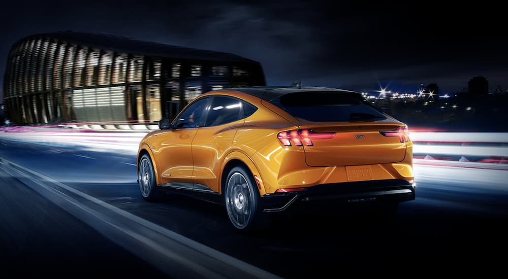 An orange 2022 Ford Mustang Mach-E is shown from the rear driving on an open road past a city at night.