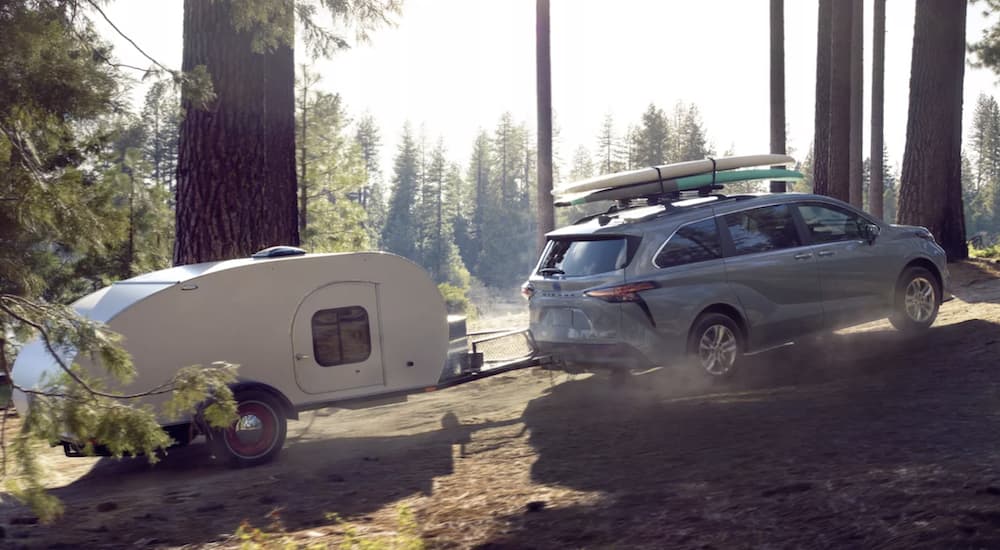A grey 2022 Toyota Sienna Woodland Edition is shown towing a small camper through the forest.