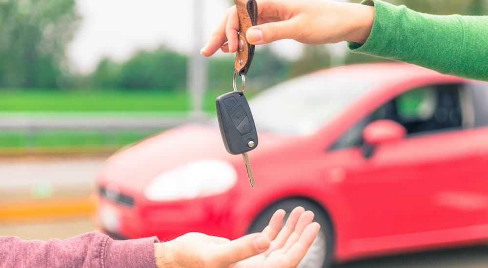 A person is shown passing a set of car keys to another person in front of a red car.