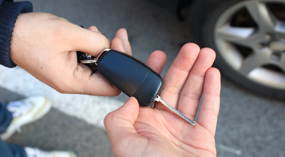 A person is passing a set of car keys to another person in front of a car.