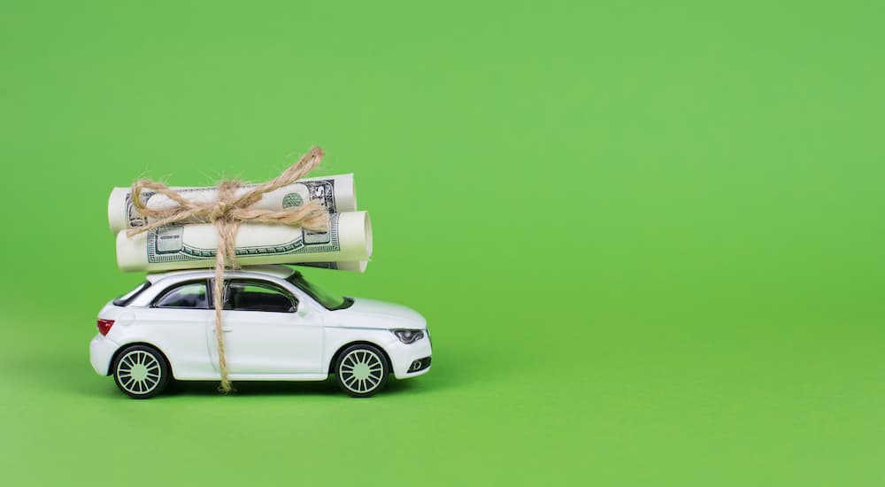 A white toy car is shown with a hundred dollar bill tied to the roof.
