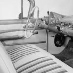 The black and white interior of a 1959 Chevy Impala shows the front seat and steering wheel.