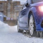 A blue car is shown idling in a snowy driveway after visiting a winter tire dealer.