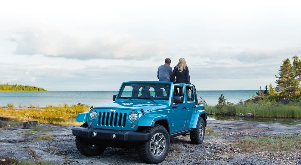 A couple is shown sitting on a blue 2021 Jeep Wrangler Rubicon near a lake.