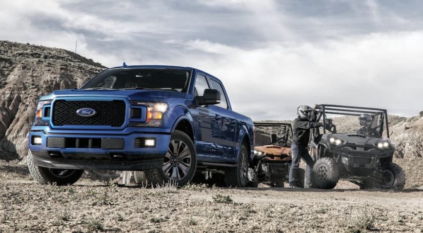 A blue 2020 Ford F-150 is shown from the front while parked next to two UTVs on a trail.