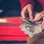 A person is shown holding a handful of cash in front of a car.