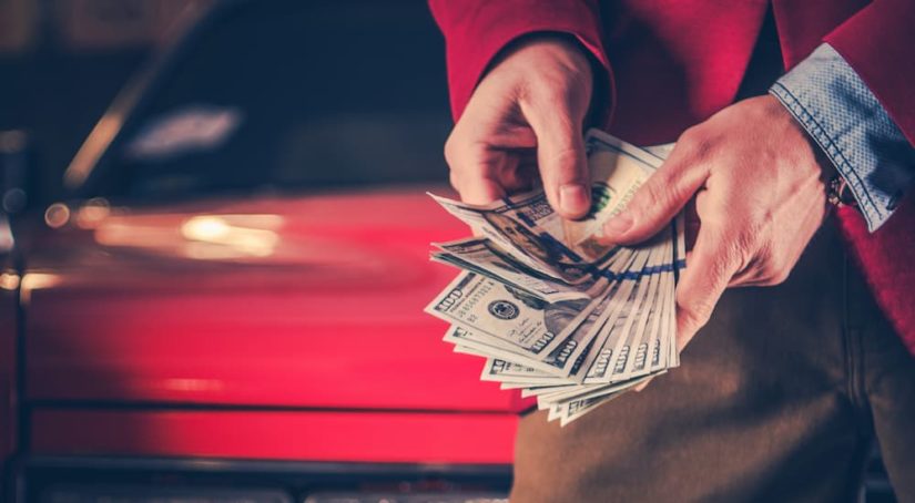 A person is shown holding a handful of cash in front of a car.