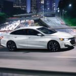 A white 2022 Chevy Malibu is shown from the side driving on a city highway.