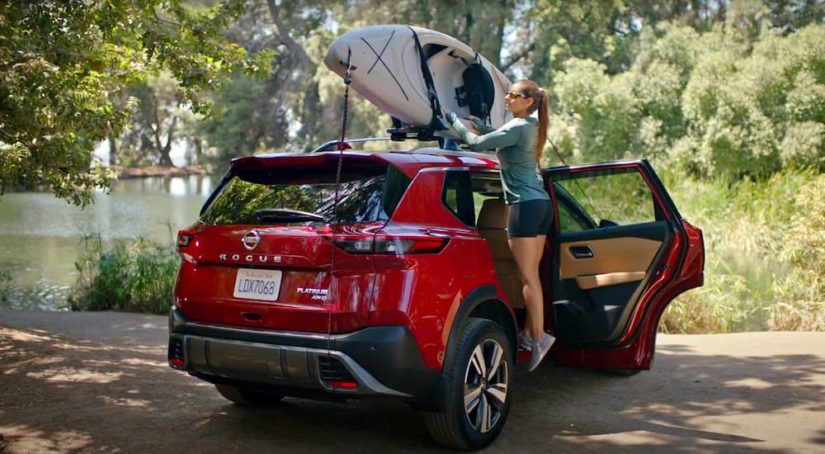 A woman is shown securing a kayak to the roof of a red 2022 Nissan Rogue Platinum.