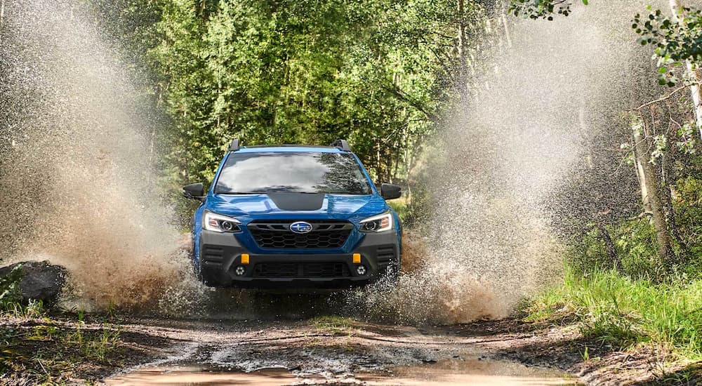 A blue 2022 Subaru Outback Wilderness is shown splashing through a mud puddle in the woods.
