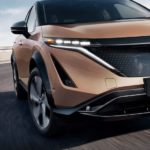A close up of a brown 2023 Nissan Ariya shows the grille and headlights.
