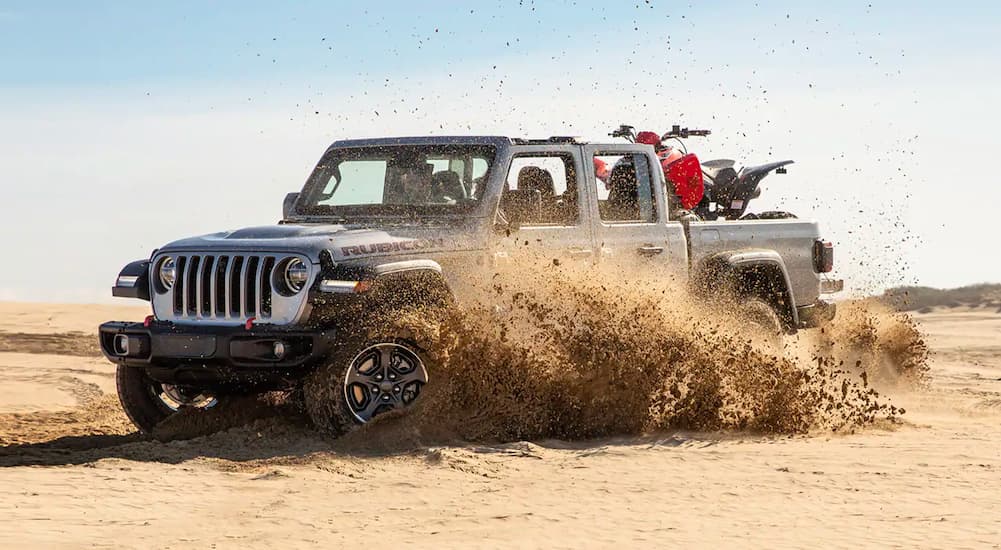 A grey 2020 Jeep Gladiator is shown kicking up sand in the desert with a 4-wheeler in the bed.