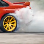 A red car is shown from the side spinning it's rear tires and kicking up smoke.