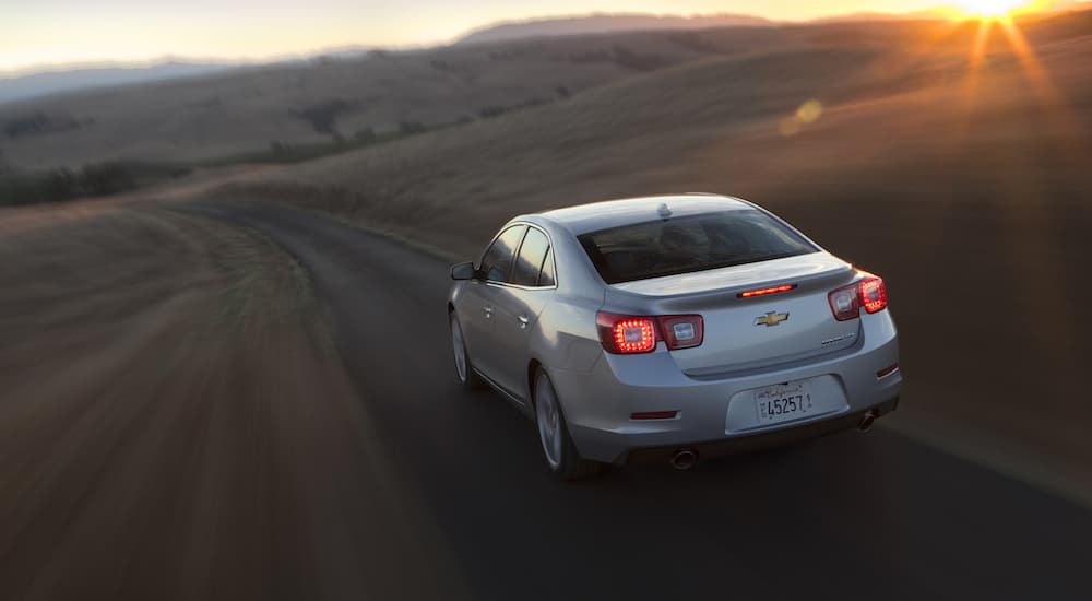 A silver 2013 Chevy Malibu Turbo is shown from the rear driving at dusk after leaving a Certified Pre-Owned Chevy dealership.
