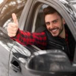 A man is shown giving a thumbs up out of the window of a grey sedan.