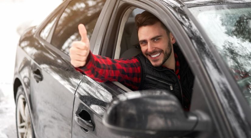 A man is shown giving a thumbs up out of the window of a grey sedan.