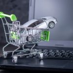 A toy car is shown in a tiny shopping cart sitting on the keyboard of a laptop.