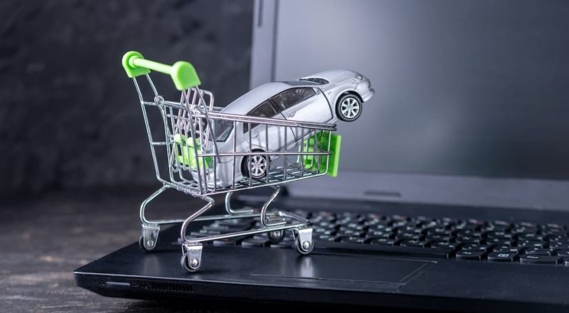 A toy car is shown in a tiny shopping cart sitting on the keyboard of a laptop.