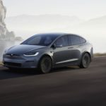 A grey Tesla Model X is shown driving on an open road near a body of water.