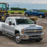 A sliver 2018 Chevy Silverado 3500 is shown from the front at an angle hooked up to a trailer at a job site.