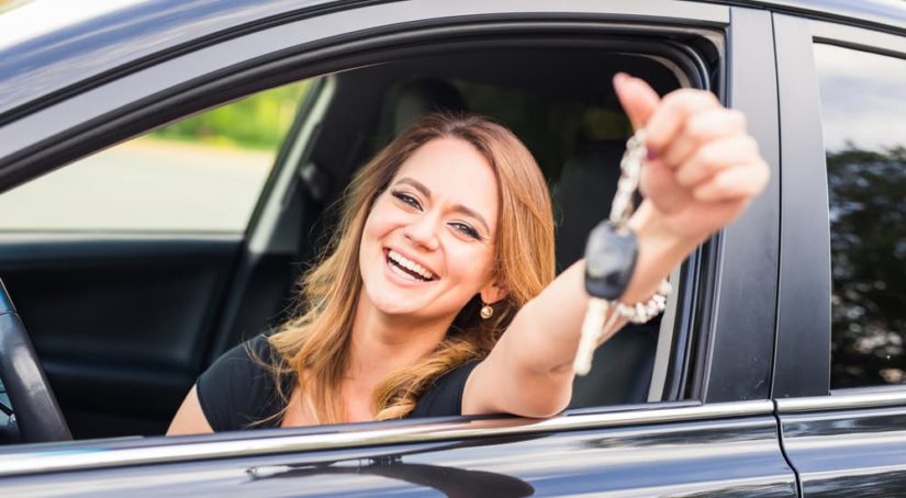 A woman is shown holding a car key out of the window of a vehicle.