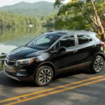 A black 2022 Buick Encore is shown from the side while it drives down a lakeside road during a 2022 Buick Encore GX vs 2022 Toyota Corolla Cross comparison.