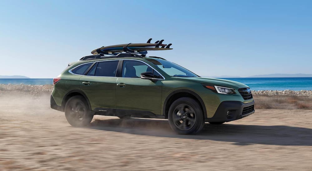 A green 2021 Subaru Outback is shown from the side as it drives down the beach with surfboards on the roof.