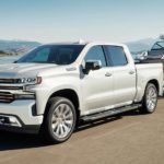 A white 2022 Chevy Silverado 1500 LTD High Country is shown towing a boat near a lake.