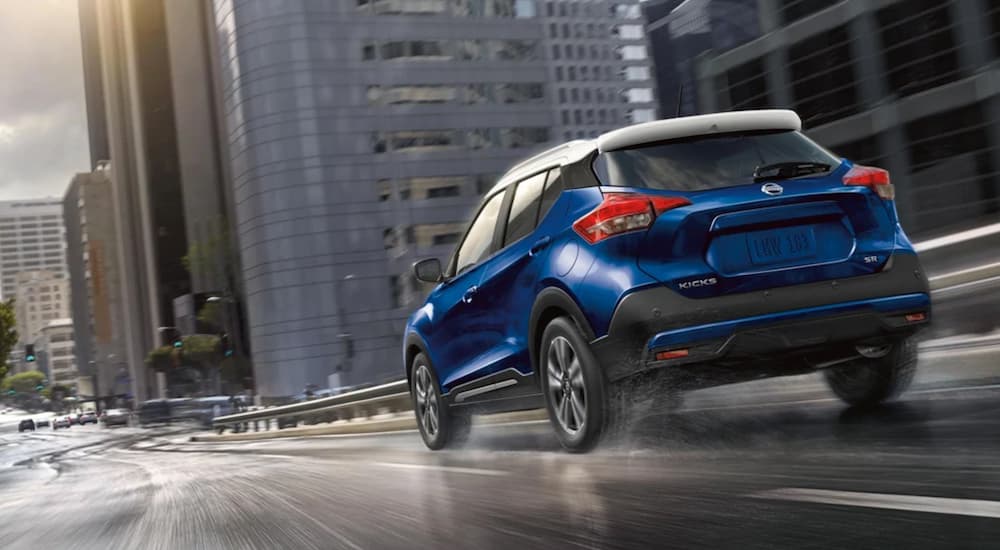 A blue 2020 Nissan Kicks is shown from the rear driving on a city highway.