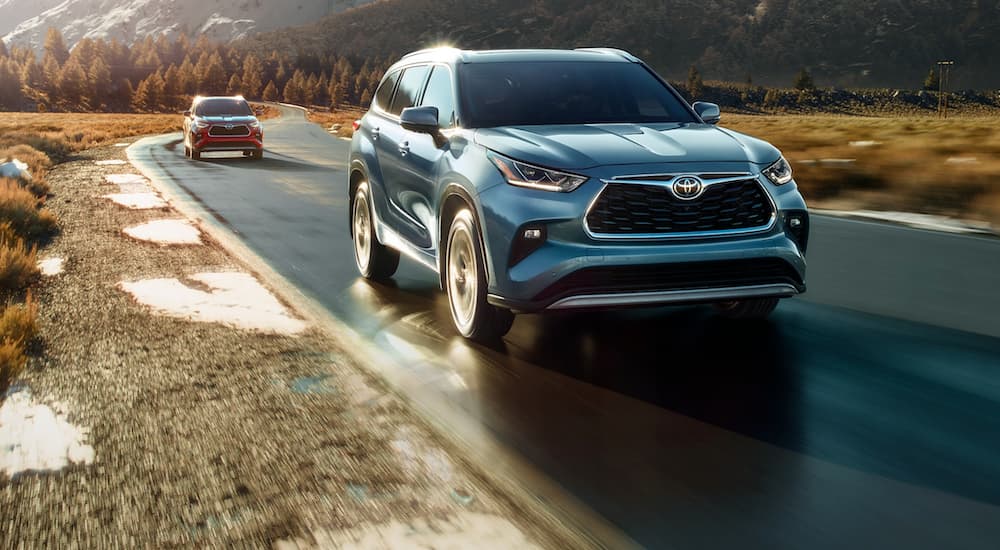 A blue 2022 Toyota Highlander is shown from the front while being followed by a red 2022 Toyota Highlander Hybrid on the road.