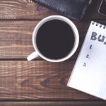 A close up shows a note pad with a budget next to a cup of coffee.