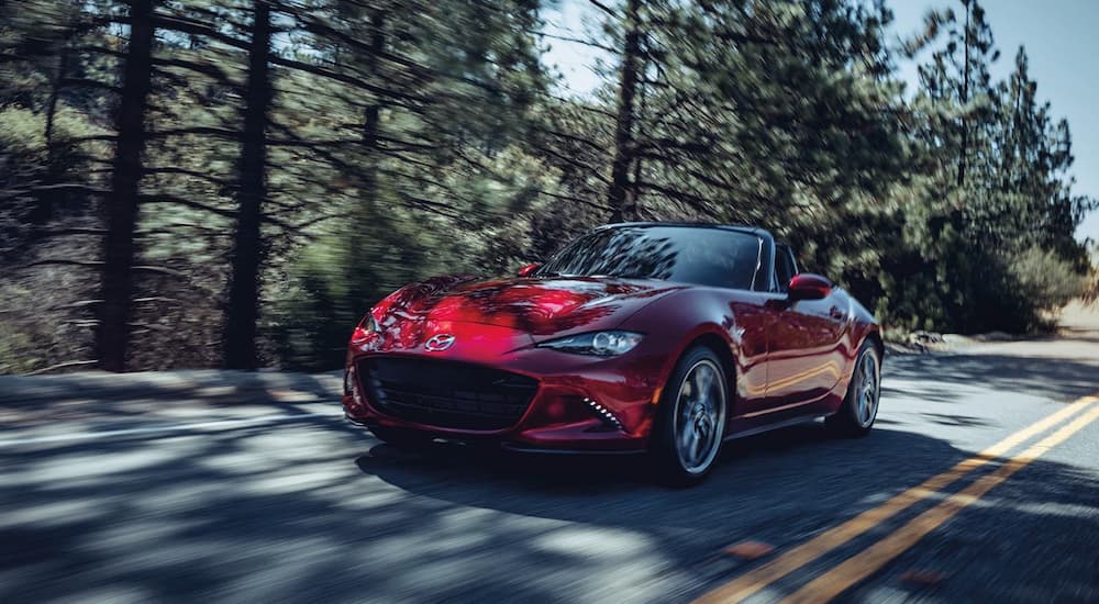 A red 2021 Mazda MX-5 Miata is shown driving down a tree-lined road.