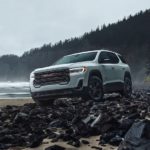 A white 2023 GMC Acadia AT4 is shown from the front at an angle on a rocky beach.