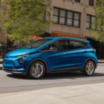 A blue 2023 Chevy Bolt EV is shown from the side driving on a city street.