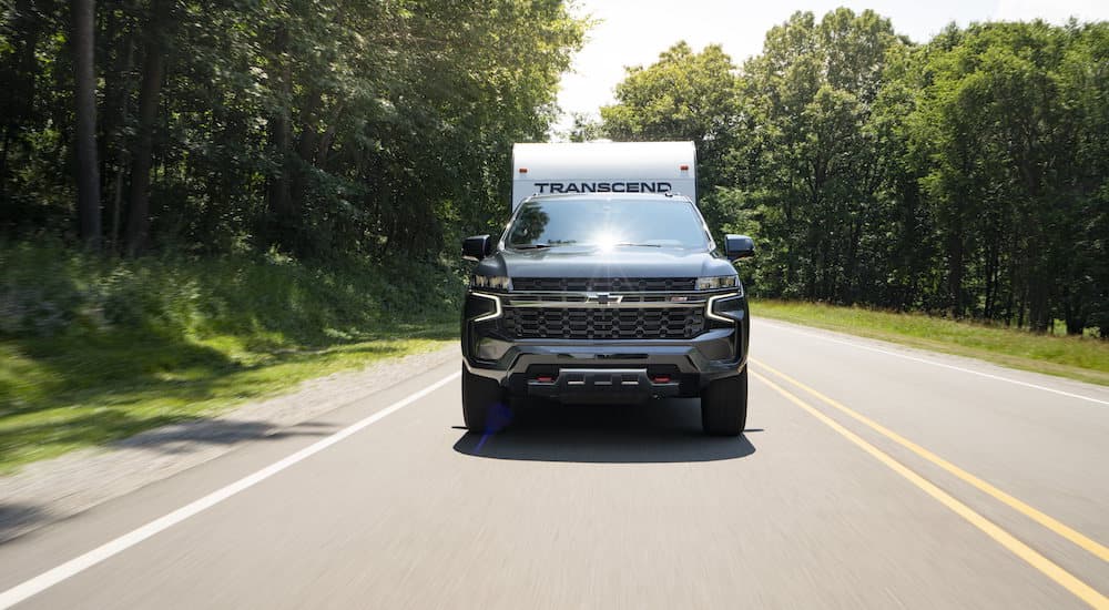 A blue 2023 Chevy Suburban Z71 is shown towing a trailer down a tree-lined road.