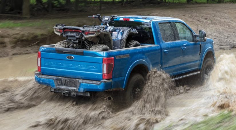 A blue 2021 Ford F-250 Super Duty Tremor is shown from the rear while driving through mud while loaded with an ATV.