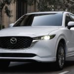 A white 2023 Mazda CX-5 is shown driving through a city.
