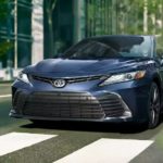 A blue 2023 Toyota Camry is shown on a city street.