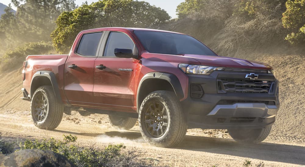 A red 2023 Chevy Colorado is shown from the side driving on a dirt road leaving a Chevy dealer.