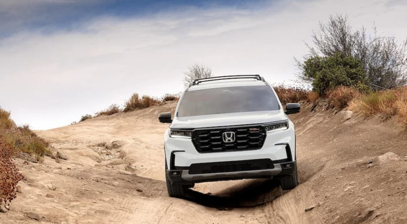 A white 2023 Honda Pilot Trailsport is shown from the front driving on a rough dirt trail.
