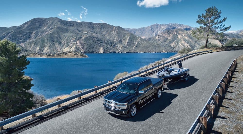 A black 2018 Chevy Silverado 1500 High Country is shown towing a boat past a lake and mountains.