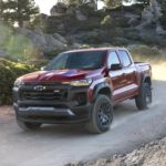 A maroon 2023 Chevy Colorado Trail Boss is shown driving on a dirt road.
