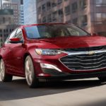 A red 2023 Chevy Malibu is shown driving on a city street.