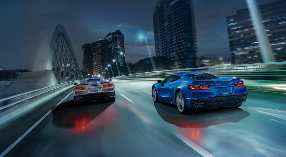 A silver and a blue 2024 Chevy Corvette E-Ray 3LZ are shown driving over a city bridge at night.
