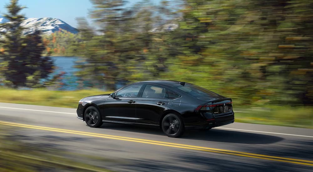 A black 2023 Honda Accord Sport-L Hybrid is shown driving past a lake and mountains.