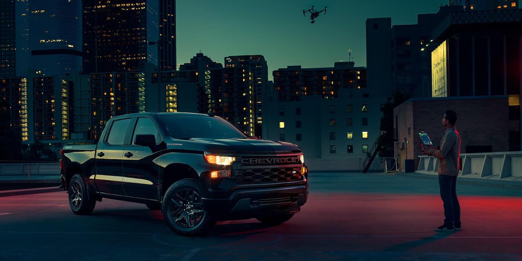 A man is shown flying a quad-rotor drone over a 2023 Chevy Silverado 1500 Custom with the Midnight Essentials package in a city parking lot at night time.
